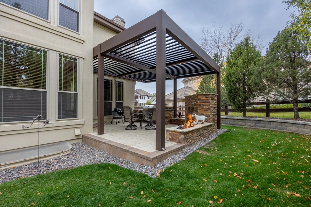 Louvered pergola over a tiled patio with a lit stone outdoor fireplace